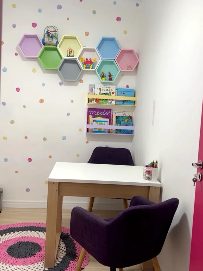 Outro ângulo da sala lúdica, com tapete de crochê colorido, poltronas e prateleiras de livros