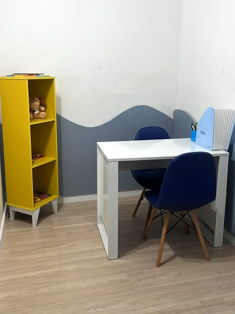 Sala de avaliação com estante amarela de brinquedos, parede em onda azul e mesa branca com cadeiras infantis